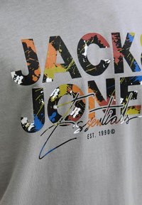 Harmaa puuvillapaita, jossa on suuri, monivärinen grafiikkateksti "JACK & JONES" roiskeen muotoisella designilla ja koukeroisella yksityiskohdalla, perustettu 1990.