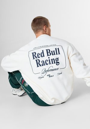 Persoon gehurkt met een wit sweatshirt met de tekst "Red Bull Racing Performance" op de rug en groene broek met witte sneakers.