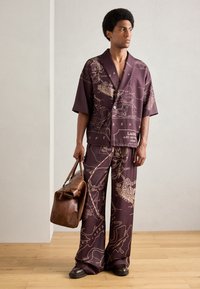 Bordeauxfarbenes Zweiteiler-Outfit mit abstraktem Kartenmuster, kurzärmeliges Hemd mit Knopfverschluss, weit geschnittene Hose und braune Ledertasche. Glatte Textur.