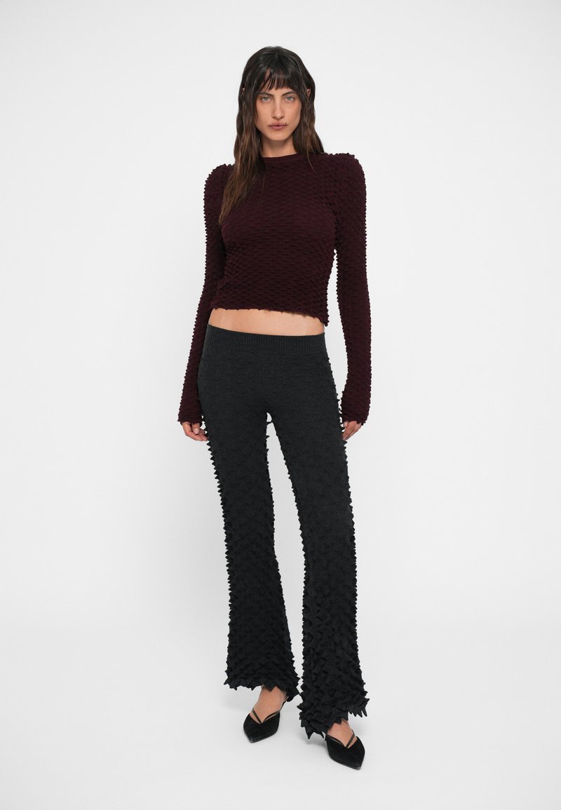 Bordeaux gekleurde, gestructureerde crop sweater met lange mouwen, gecombineerd met zwarte flare broeken die een vergelijkbaar gestructureerd patroon en herkenbare franjes aan de onderkant hebben.