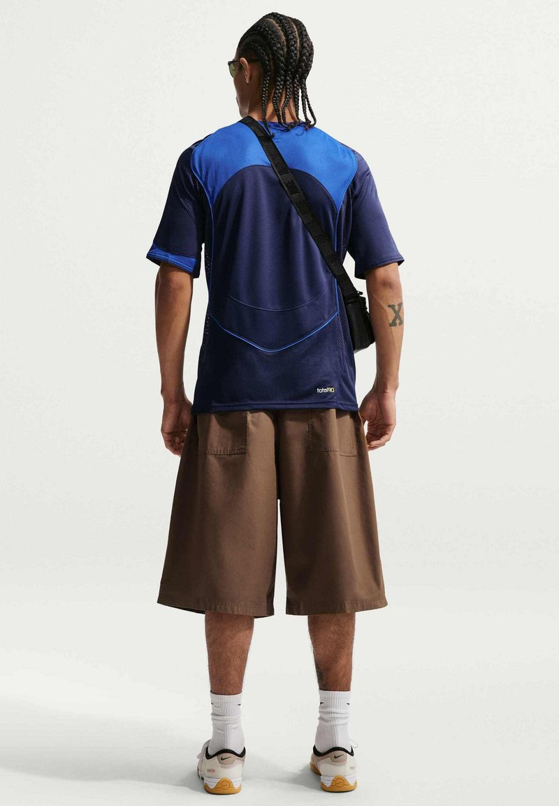 Hombre con trenzas que lleva una camisa azul marino y azul, pantalones cortos marrones anchos, calcetines blancos, zapatillas deportivas y un bolso negro cruzado, de espaldas.