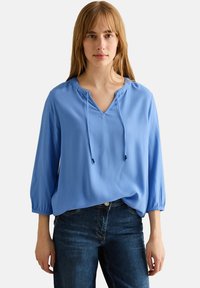 Blouse légère bleue avec un décolleté en V, un détail de cordon de serrage et des manches trois-quarts. Associée à un jean en denim noir, présentant une coupe décontractée.