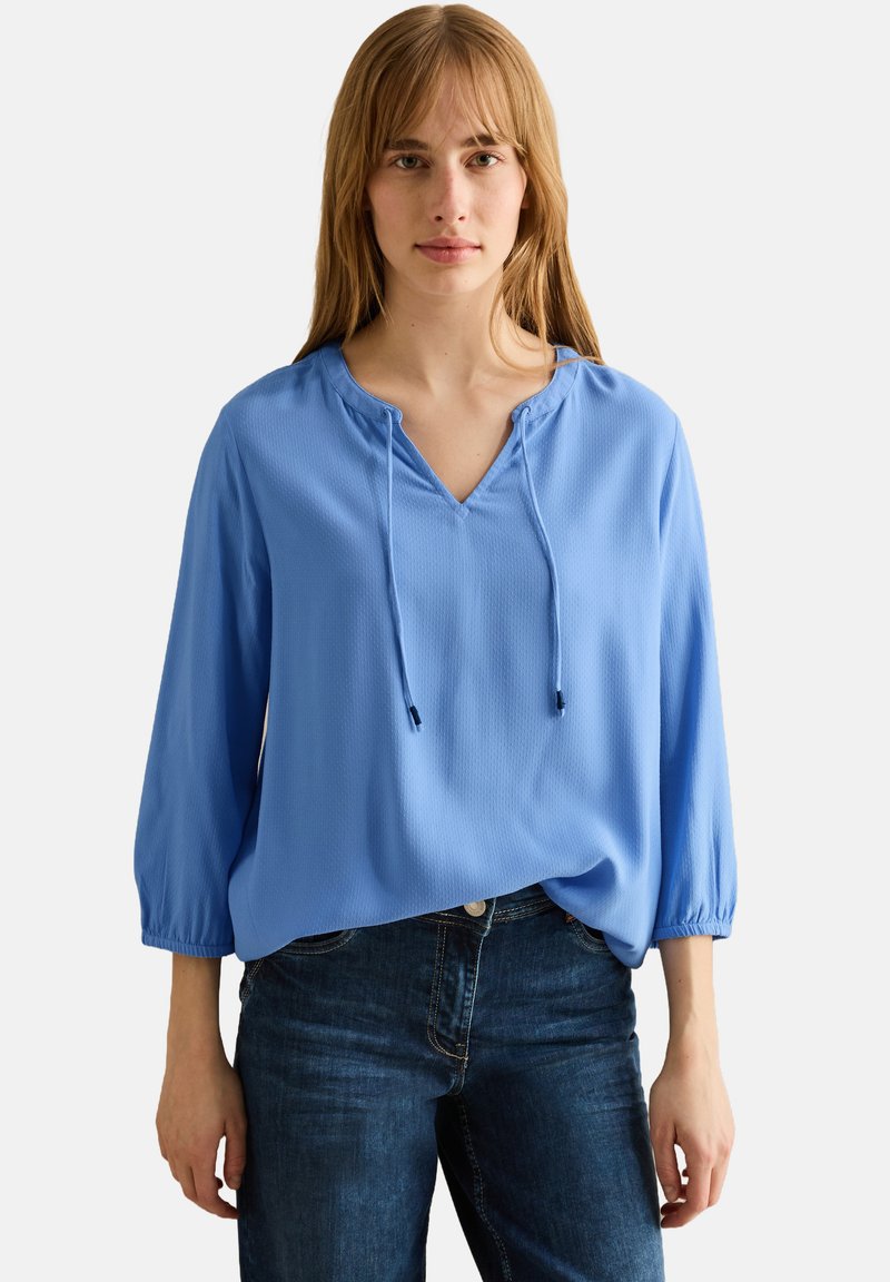 Blouse légère bleue avec un décolleté en V, un détail de cordon de serrage et des manches trois-quarts. Associée à un jean en denim noir, présentant une coupe décontractée.