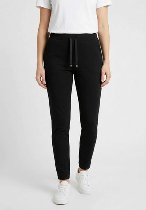 Persona che indossa pantaloni jogger neri aderenti con coulisse in vita, t-shirt bianca e sneakers bianche, in piedi davanti a uno sfondo chiaro semplice.