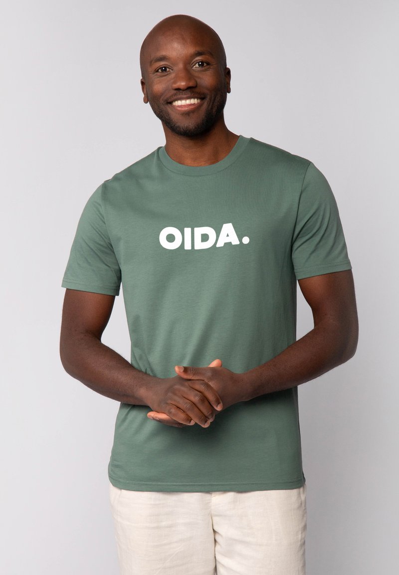 Grünes Baumwoll-T-Shirt mit dem weißen Text "OIDA." Kurze Ärmel, Rundhalsausschnitt, glatte Textur, getragen mit hellfarbigen Hosen.