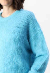 Pull tricoté bleu clair avec une texture douce et duveteuse, col rond et coupe légèrement oversize. Comprend des manches longues et amples.