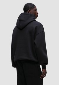 Černá mikina s kapucí a volným střihem, vyrobená z měkkého materiálu. Má spadlé ramena a žebrované manžety. Zobrazený zadní pohled.