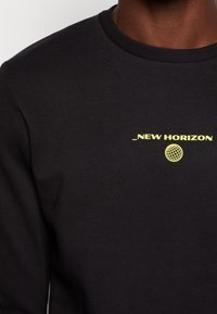Svart sweatshirt med gul text "_NEW HORIZON" och en globgrafik. Rund hals, texturerad tyg, visas från bröstet och uppåt.
