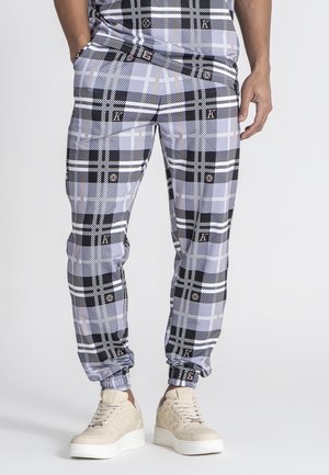 Grau-schwarze Karotten-Hosen mit elastischen Bündchen, die eine glatte Textur und Logo-Details aufweisen. Kombiniert mit hellen Sneakern.