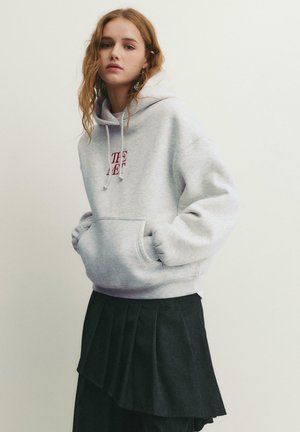 Tânără cu păr ondulat, purtând un hanorac gri deschis cu text roșu și o fustă plisată neagră stratificată, stând pe un fundal simplu.