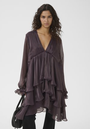 Femme aux longs cheveux bouclés portant une robe superposée, transparente de couleur violet foncé, avec manches longues, tenant un sac à main noir sur un fond uni.