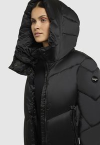 Schwarze Pufferjacke mit einer großen, dicken Kapuze, glatter Textur, Druckknopf- und Reißverschluss, und einem Emblem am linken Ärmel. Entworfen mit einer kastigen Form.