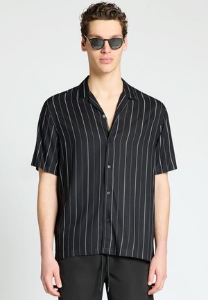 Uomo con camicia nera a maniche corte con righe verticali bianche e occhiali da sole scuri, in piedi davanti a uno sfondo uniforme.