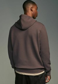 Brun hætte sweatshirt med et struktureret, quiltet mønster. Har en hætte med snoretræk og ribbede manchetter, lagdelt over en lysefarvet top.