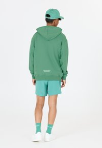 Grüner Kapuzenpullover mit bequemer Passform, kombiniert mit passenden tealfarbenen Shorts. Weiße Sneaker und tealfarbene Socken sind sichtbar, mit dezenten Branding-Details.