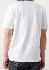 Bílý bavlněný tričko s krátkými rukávy, kulatým výstřihem a hladkou texturou, viděné ze zadní strany, v kombinaci s vzorovanými šortkami.