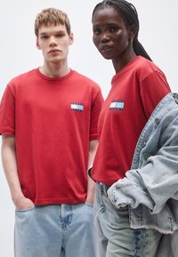 Duas pessoas vestem t-shirts vermelhas iguais com um pequeno logótipo ao peito. O modelo masculino combina a sua t-shirt com jeans azul claro, enquanto a modelo feminina usa uma jaqueta de ganga por cima da sua.