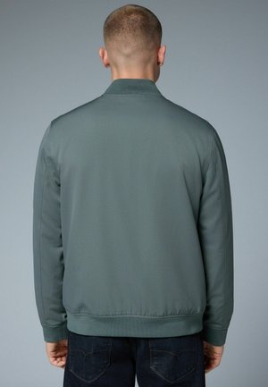 Giacca bomber grigia con una texture liscia e opaca, colletto e polsini a coste. Presenta una fascia in vita aderente, vista dal retro. Jeans scuri visibili.