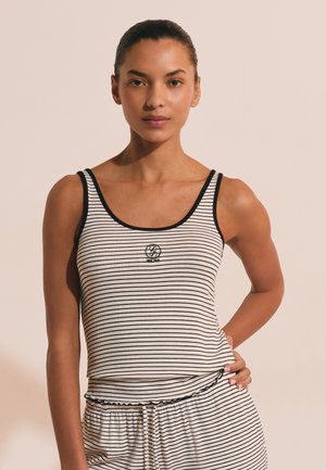 Frau mit zurückgebundenem Haar trägt ein schwarz-weiß gestreiftes Tanktop und passende Hose, steht mit einer Hand an der Hüfte.