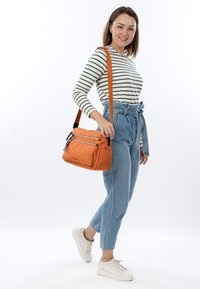Smiling woman wearing striped long-sleeve shirt, high-waisted jeans, and white sneakers, carrying orange crossbody bag on shoulder.