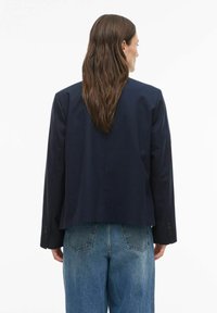 Femme aux longs cheveux bruns portant un blazer bleu marine et un jean bleu, debout de dos sur un fond blanc uni.