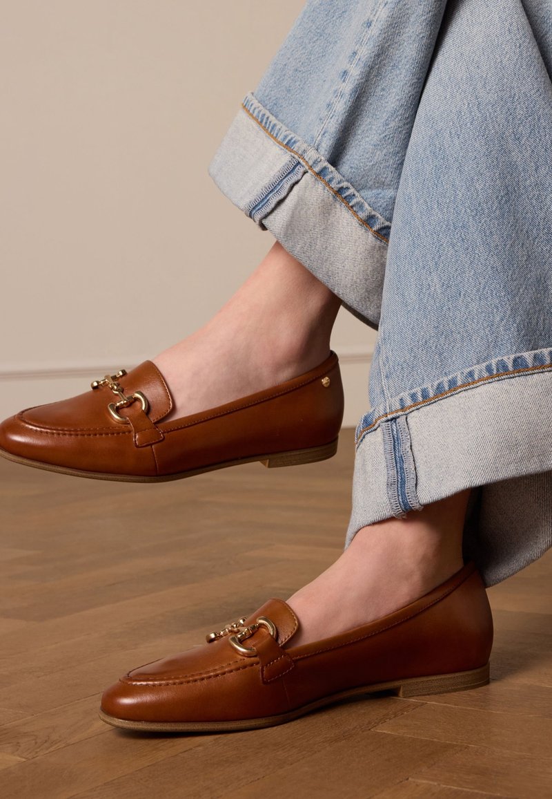 Chevilles fines portant un jean bleu clair avec revers et des mocassins en cuir marron avec des détails en métal doré sur un parquet en bois