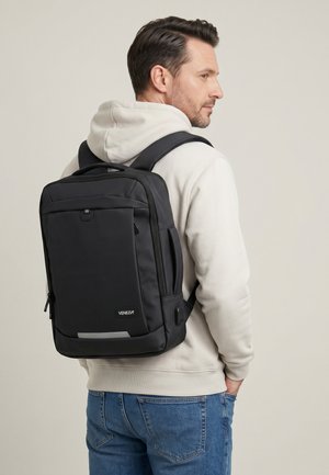 Mann trägt hellbeigen Hoodie und blaue Jeans mit schwarzem rechteckigem Venezia-Rucksack, seitlich vor eintönigem Hintergrund.