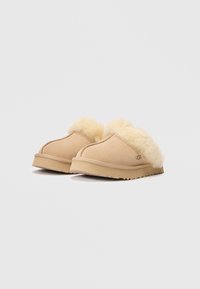 Beige semskede tøfler med en deilig kremfarget ullfore, et buet design og en flat gummisåle med et strukturert mønster.