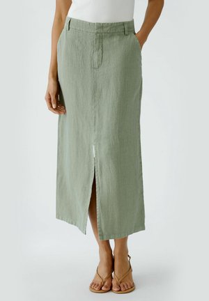 Kobieta ubrana w jasnozieloną lnianą spódnicę midi z przednim rozcięciem, biały top bez rękawów oraz beżowe sandały na paski, stojąca na neutralnym tle.