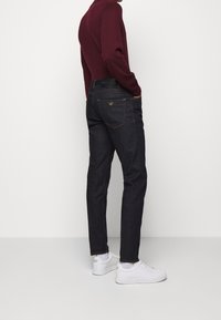 Man som bär mörkblå jeans, vita sneakers och en vinröd långärmad skjorta, står med händerna i fickorna mot en enfärgad bakgrund.