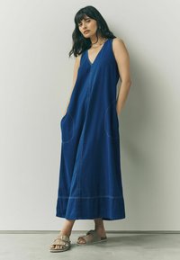 Rochie din denim albastru, cu decolteu în V, fără mâneci, cu buzunare și o siluetă lungă, fluidă. Prezintă cusături contrastante și accente minimaliste.