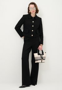 Schwarze maßgeschneiderte Jacke mit goldenen Knöpfen, kombiniert mit weit geschnittenen schwarzen Hosen. Ergänzt mit einer gestreiften Handtasche in Beige und Schwarz, High Heels und Sonnenbrille.