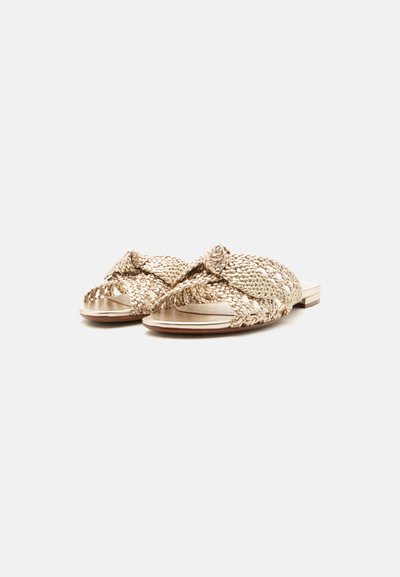 Επίπεδα σανδάλια slip-on με φαρδιούς πλεγμένους μπεζ ιμάντες που διασταυρώνονται πάνω από τα ακάλυπτα δάχτυλα, επίπεδη σόλα και ελαφριά φτέρνα, εμφανίζονται σε λευκό φόντο.