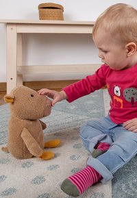 Tout-petit en chemise rouge et chaussettes rayées assis sur un tapis, touchant doucement le nez d'un ours en peluche marron.