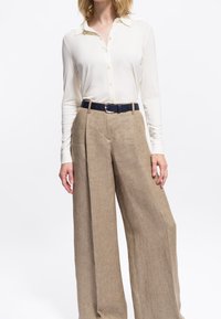 Donna che indossa una camicetta crema con bottoni infilata in pantaloni beige a vita alta e larghi con una cintura nera, in piedi contro uno sfondo bianco.