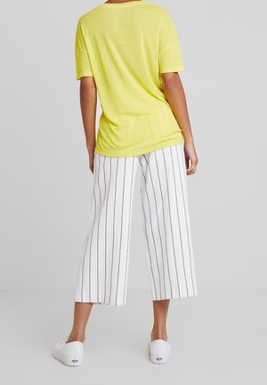 Persoon staand, draagt een losse gele top met korte mouwen, witte wijde broek met zwarte verticale strepen en witte sneakers, tegen een effen achtergrond.