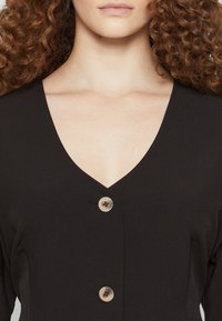 Μαύρο φόρεμα από ύφασμα με βαθύ V-ντεκολτέ, με τρία στρογγυλά κουμπιά από αντίθετο υλικό στη μέση, με απαλή υφή.