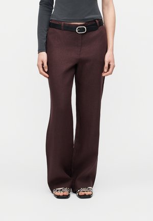 Personne portant un pantalon large brun foncé avec une ceinture noire, un haut à manches longues gris, et des sandales noires à lanières imprimé serpent.