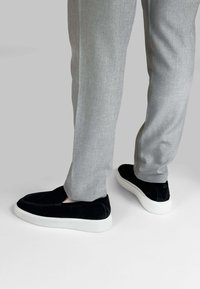 Zwarte suède instapschoenen met witte rubberen zolen, gecombineerd met lichtgrijze gestructureerde broeken, die een modern, minimalistisch ontwerp laten zien.