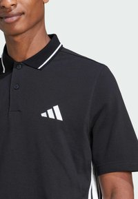 Sort polo-shirt lavet af struktureret stof, med en hvid krave-stribe, to knapper og et hvidt logo med tre striber på venstre bryst.