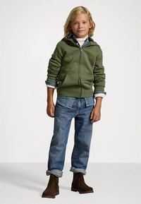 Πράσινη ζακέτα με φερμουάρ, μπλε τζιν παντελόνι, καφέ μπότες. Η ζακέτα έχει δύο μπροστινές τσέπες και λεπτομέρειες από ριμπ στις μανσέτες. Το μοντέλο έχει ατημέλητα ξανθά μαλλιά.