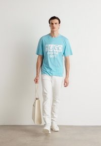 Lyseblå bomulls t-skjorte med hvit grafisk tekst "HUGO CREATIVE DEPT.", kombinert med hvite jeans, og bærer en beige totebag.