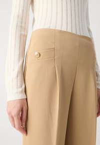 Beige skræddersyede bukser lavet af tekstureret stof, med en sidelomme og en guldknap som accent. Båret med en hvid ribbet sweater.