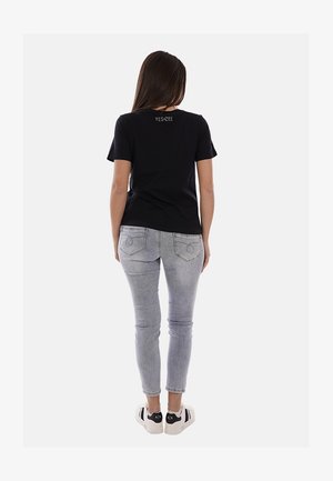 Mujer con camiseta negra de manga corta con el texto "YES ZEE" en la parte trasera, jeans gris claro y zapatillas blancas con detalles en negro, de pie.
