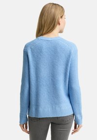 Frau mit langen blonden Haaren, die einen hellblauen Strickpullover und graue Jeans trägt, mit dem Gesicht abgewandt vor einem schlichten weißen Hintergrund.