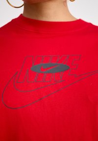 Raudonas medvilnės marškinėliai su išsiskiriančiu pilku Nike logotipu ir swoosh dizainu, su apvaliu iškirpimu ir trumpomis rankovėmis. Lygus paviršius.
