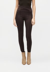 Γυναίκα που φοράει παντελόνι ψηλόμεσο, στενή εφαρμογή σε σκούρο καφέ χρώμα με τσεπάκια με κουμπιά, συνδυασμένο με ταμπά σουέτ μποτάκια μέχρι τον αστράγαλο, σε λευκό φόντο.