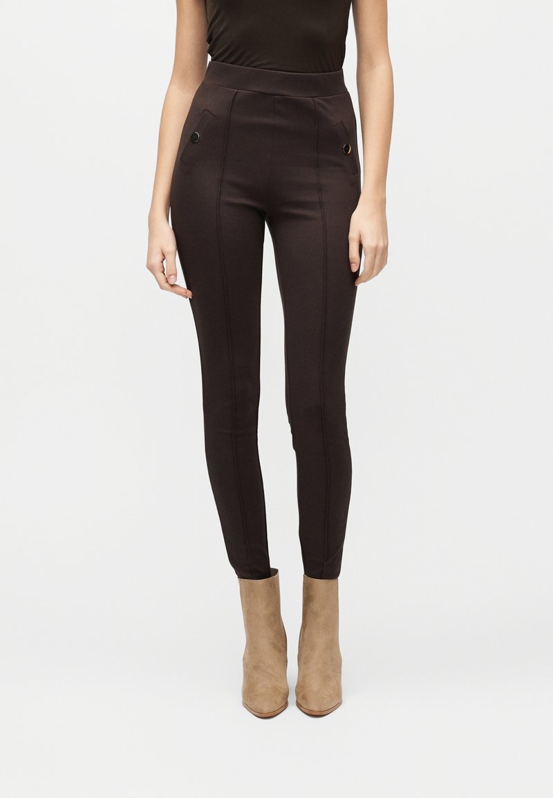 Γυναίκα που φοράει παντελόνι ψηλόμεσο, στενή εφαρμογή σε σκούρο καφέ χρώμα με τσεπάκια με κουμπιά, συνδυασμένο με ταμπά σουέτ μποτάκια μέχρι τον αστράγαλο, σε λευκό φόντο.