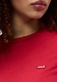 Κλείσιμο σε άτομο που φορά ει κόκκινο t-shirt Levi's, εμφανίζοντας τα χείλη, το πηγούνι, το λαιμό, τα σγουρά μαλλιά και το λογότυπο Levi's στο στήθος.