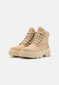 Bottes chevilles en suède beige avec bout rond, œillets en métal et lacets crème, dotées d'une semelle en caoutchouc texturée et d'un logo sur le côté.